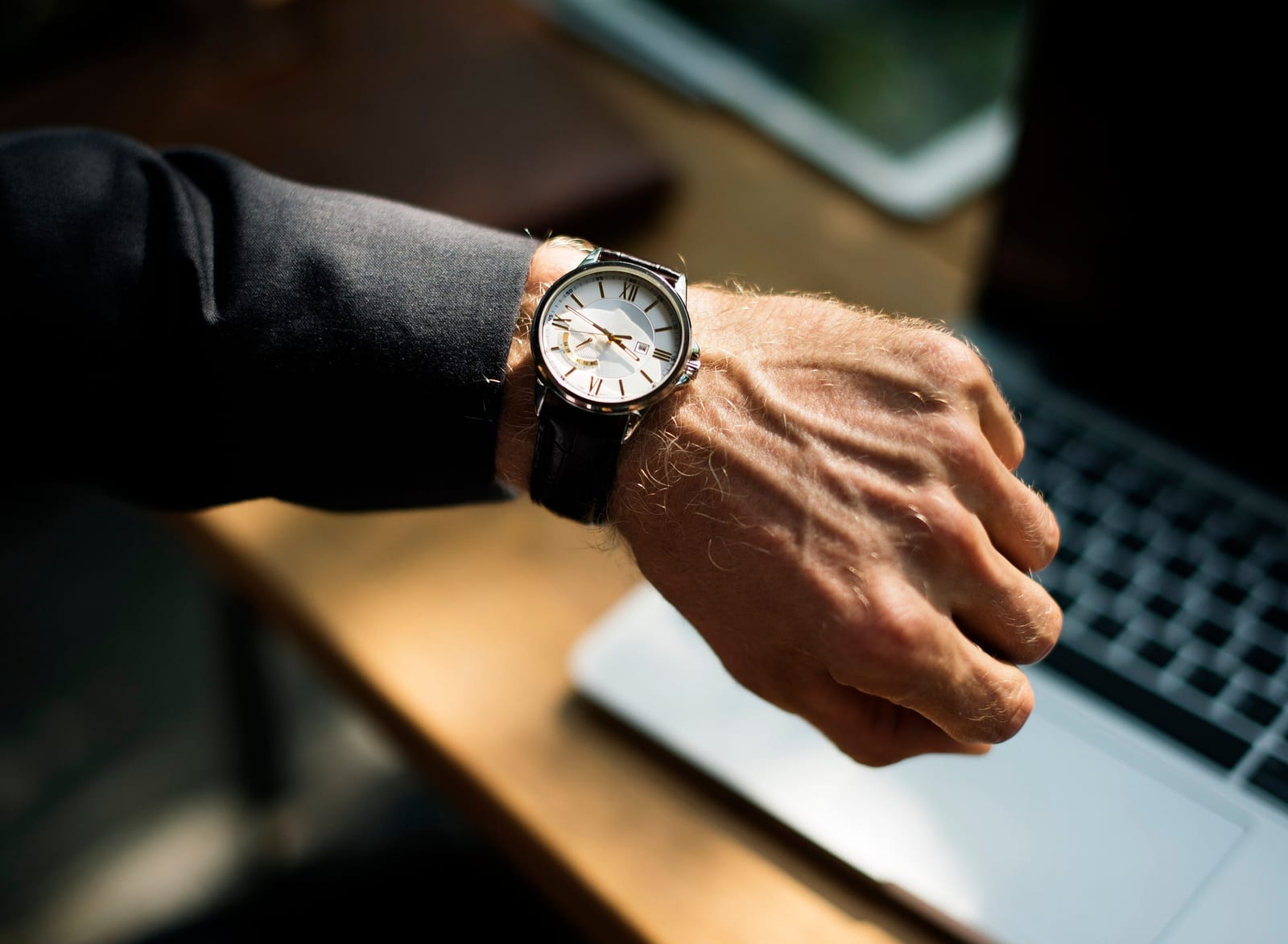 A wristwatch with a white dial on a wrist, beside a laptop