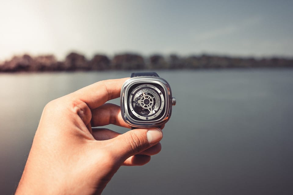 SEVENFRIDAY automatic watch with an open dial, held up against a lake