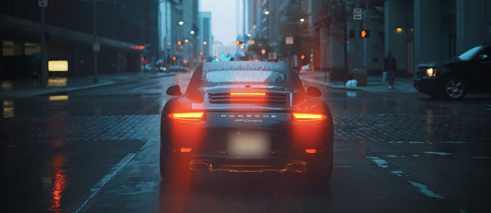 Porsche 911 Carrera from behind on a wet city street at dusk, tail lights glowing